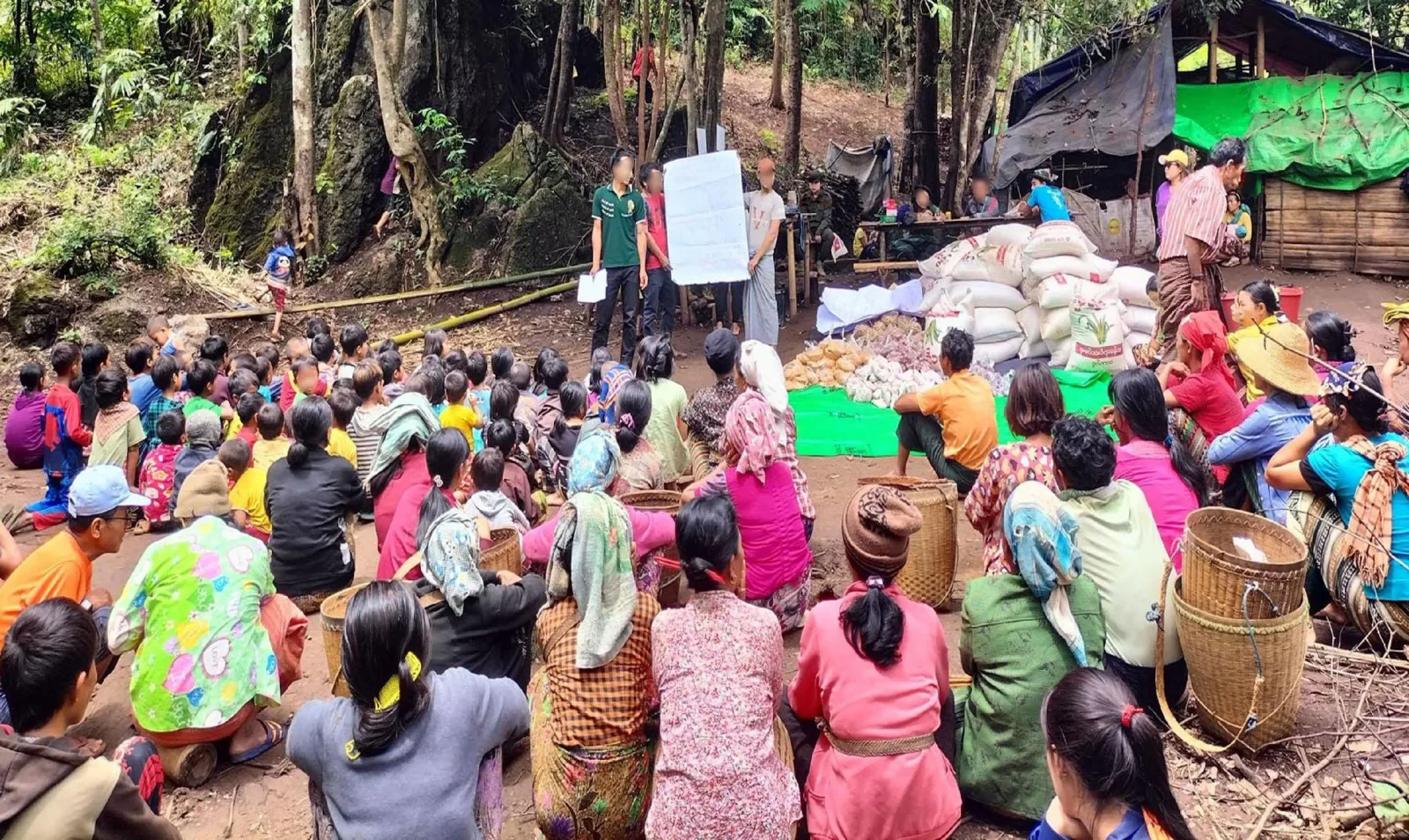 Families sit together as a distribution is explained beside a stack of rice sacks at a displacement site