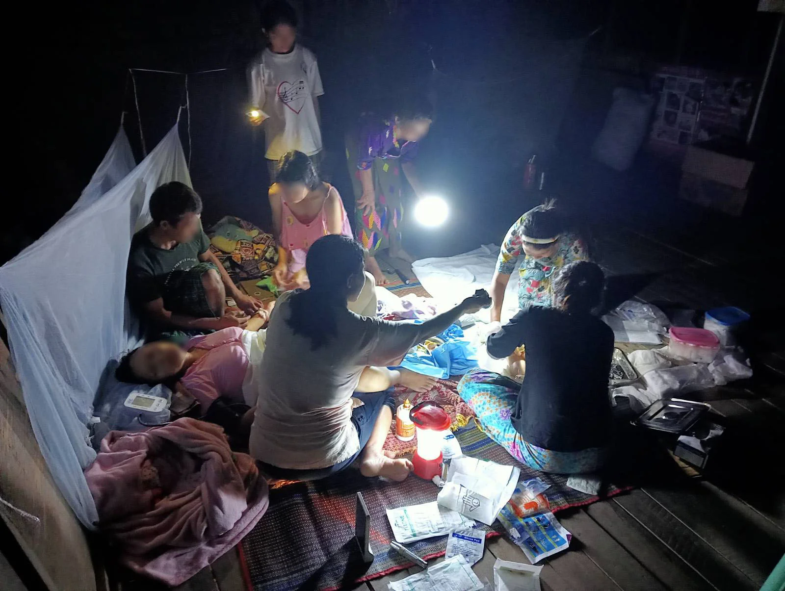 A health team treats a patient by lantern light on a mat inside a village house at night