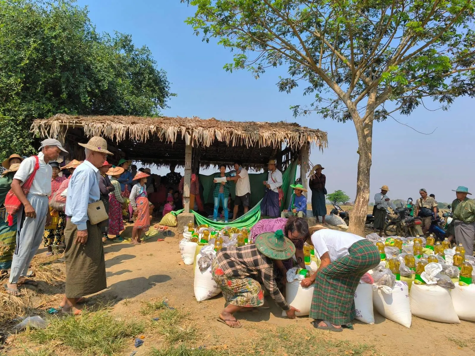 Families queue beside sacks of rice and bottles of cooking oil at a distribution point in Bago Region