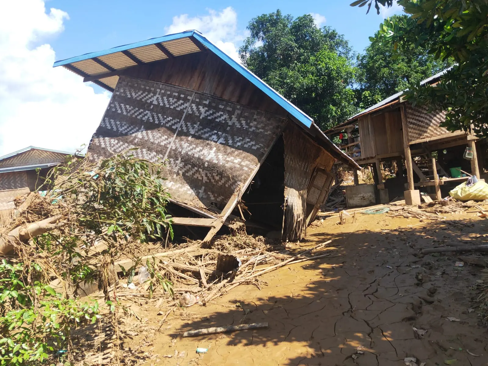 A bamboo house pushed off its posts by floodwater, surrounded by dried mud and debris